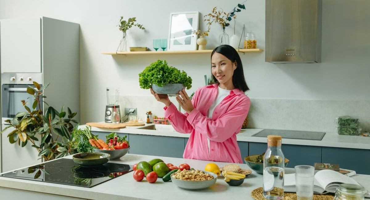 Woman in a kitchen holding fresh lettuce, surrounded by healthy ingredients like tomatoes and avocados, promoting healthy eating.