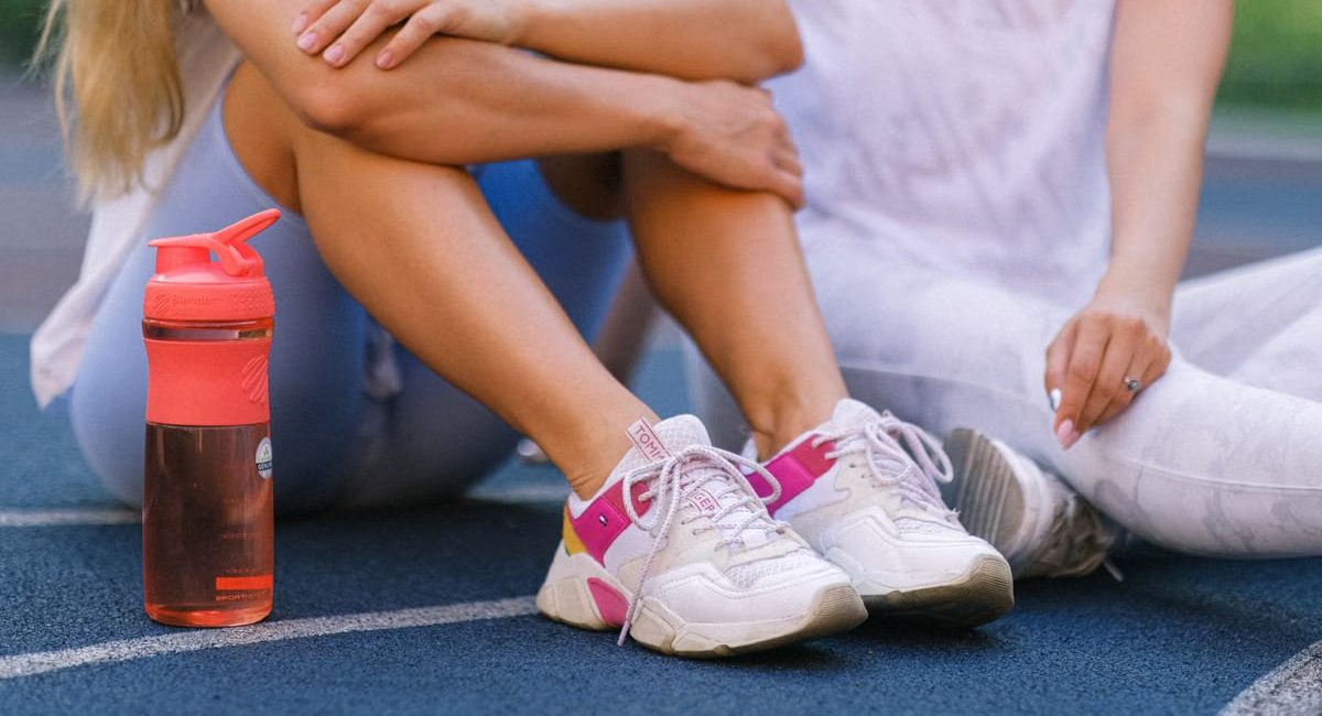 Crop anonymous female friends in activewear sitting on sports ground after training near bottle with water in daytime