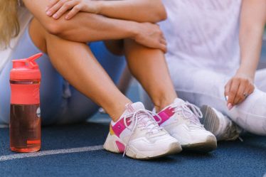 Crop anonymous female friends in activewear sitting on sports ground after training near bottle with water in daytime